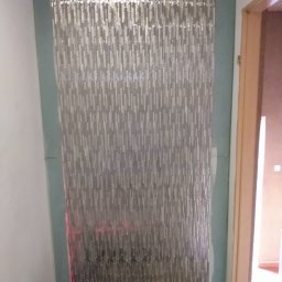 Zdzisław Hamerski - Ściana wyłożona mozaiką w odcieniach szarości, z widocznymi niedociągnięciami przy krawędziach i resztkami zaprawy, oświetlona laserowym poziomem, na tle fragmentu drzwi.