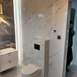 Zdzisław Hamerski - Nowoczesna łazienka z białymi płytkami imitującymi marmur z brązowymi żyłkami, podwieszana toaleta i biała szafka z szufladami, oświetlenie LED w pionowej wnęce, widoczny fragment osoby w przejściu.