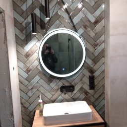 Zdzisław Hamerski - Ukończona łazienka z umywalką nablatową, lustrem z podświetleniem LED i wzorem jodełki na ścianie z płytek, widoczne lampy wiszące i silikon sanitarny.