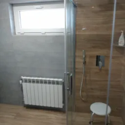 Nowoczesna łazienka z kabiną prysznicową typu walk-in, wyłożona szarymi płytkami na ścianach i drewnopodobnymi płytkami na podłodze oraz wewnątrz kabiny. Biały grzejnik pod oknem i małe siedzisko...