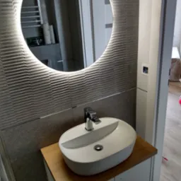 Nowoczesna łazienka z umywalką nablatową i okrągłym lustrem z podświetleniem LED, ściana wyłożona płytkami imitującymi fale.