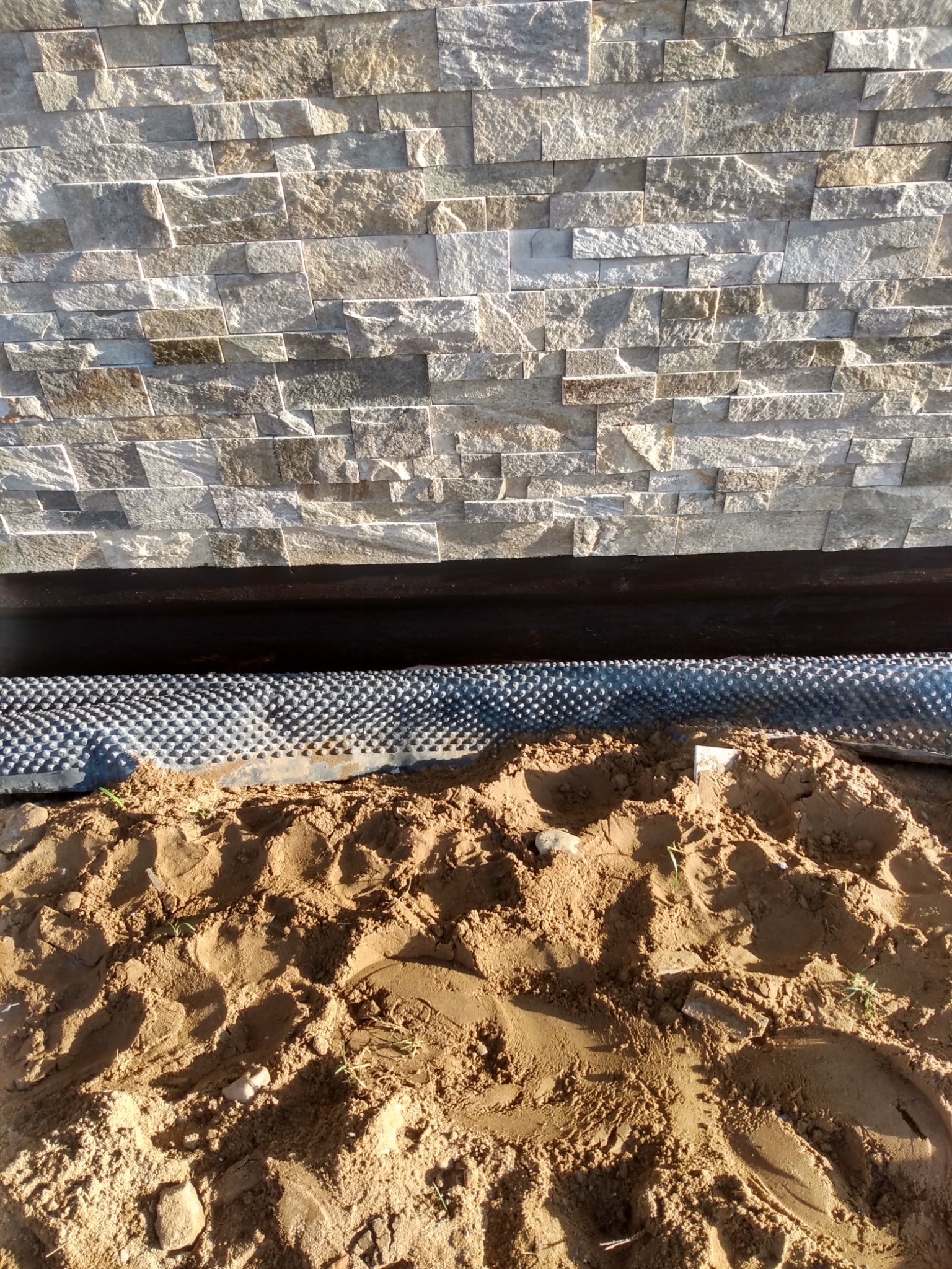 Ściana wyłożona dekoracyjnym kamieniem elewacyjnym, widoczna czarna izolacja przeciwwilgociowa oraz folia kubełkowa i piasek w wykopie.