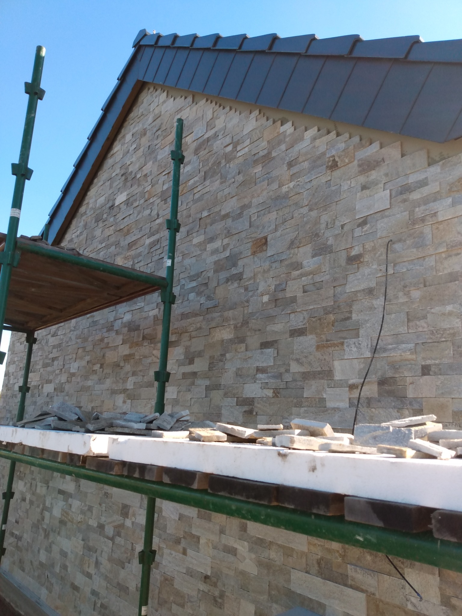 Elewacja domu w trakcie wykańczania kamieniem dekoracyjnym, widoczne rusztowanie i fragment dachu z ciemnej dachówki.