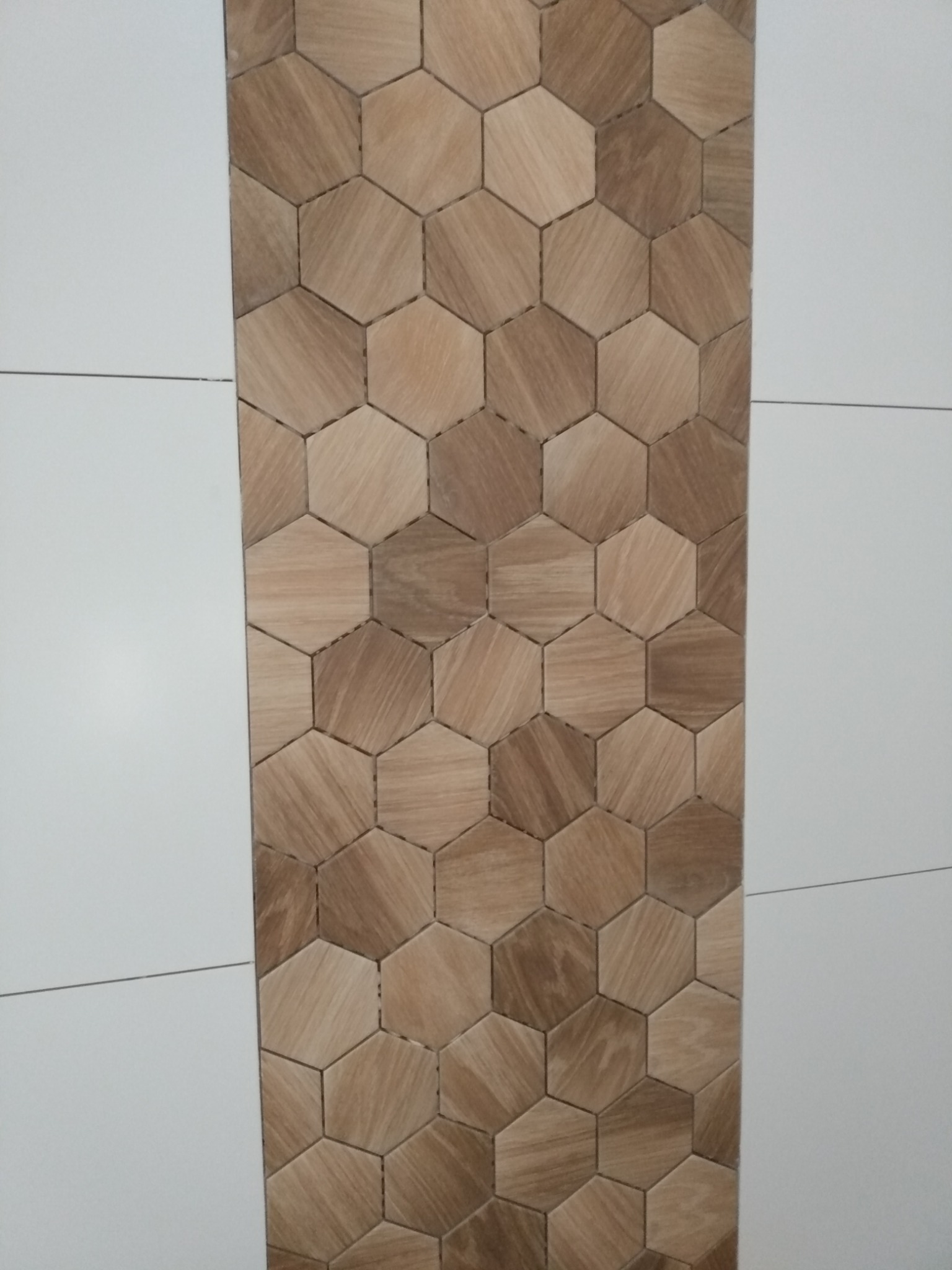 Fragment ściany z heksagonalnymi płytkami w odcieniach brązu, imitującymi drewno, sąsiadującymi z białymi, prostokątnymi płytkami.