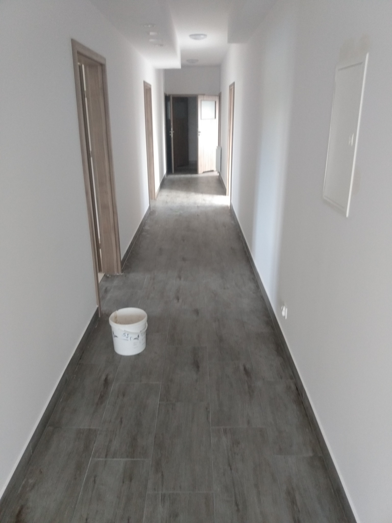 Długi korytarz po remoncie z szarymi panelami podłogowymi imitującymi drewno, pomalowanymi na biało ścianami i widocznym wiadrem po farbie.