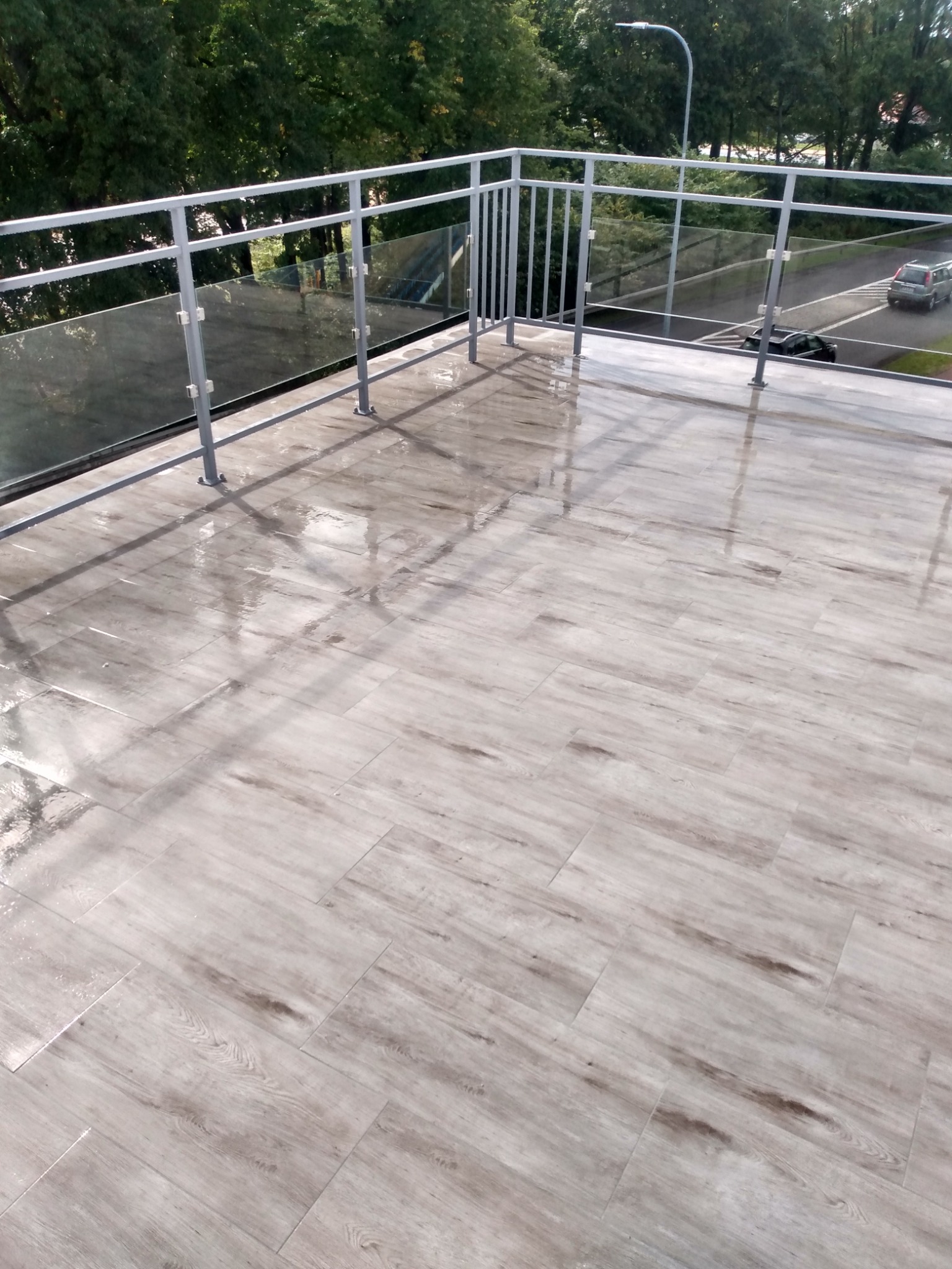 Świeżo ułożona, jasna, drewnopodobna terakota na balkonie z metalową balustradą i szklanymi panelami, mokra od deszczu, odbijająca światło.
