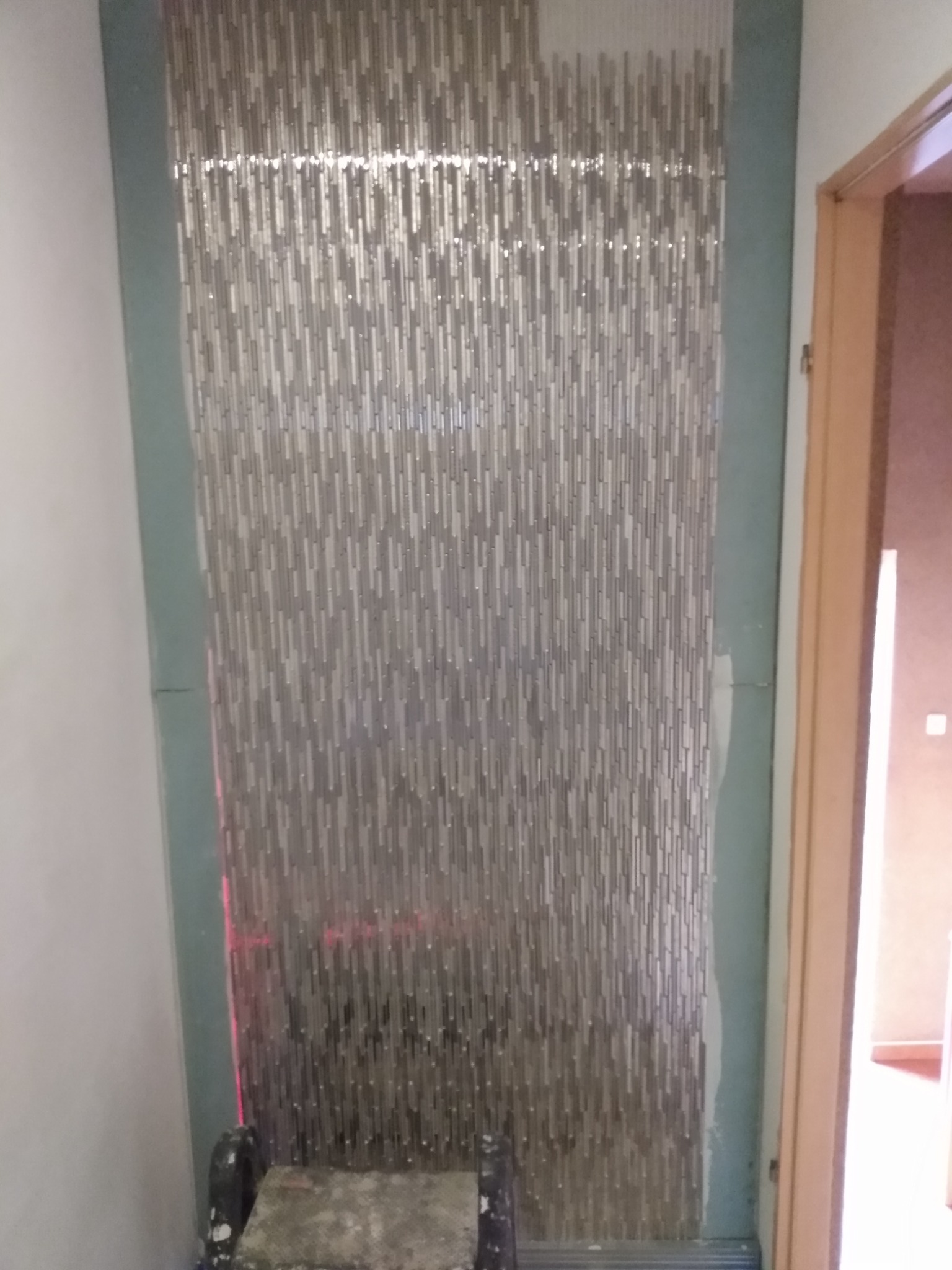 Ściana wyłożona mozaiką w odcieniach szarości, z widocznymi niedociągnięciami przy krawędziach i resztkami zaprawy, oświetlona laserowym poziomem, na tle fragmentu drzwi.