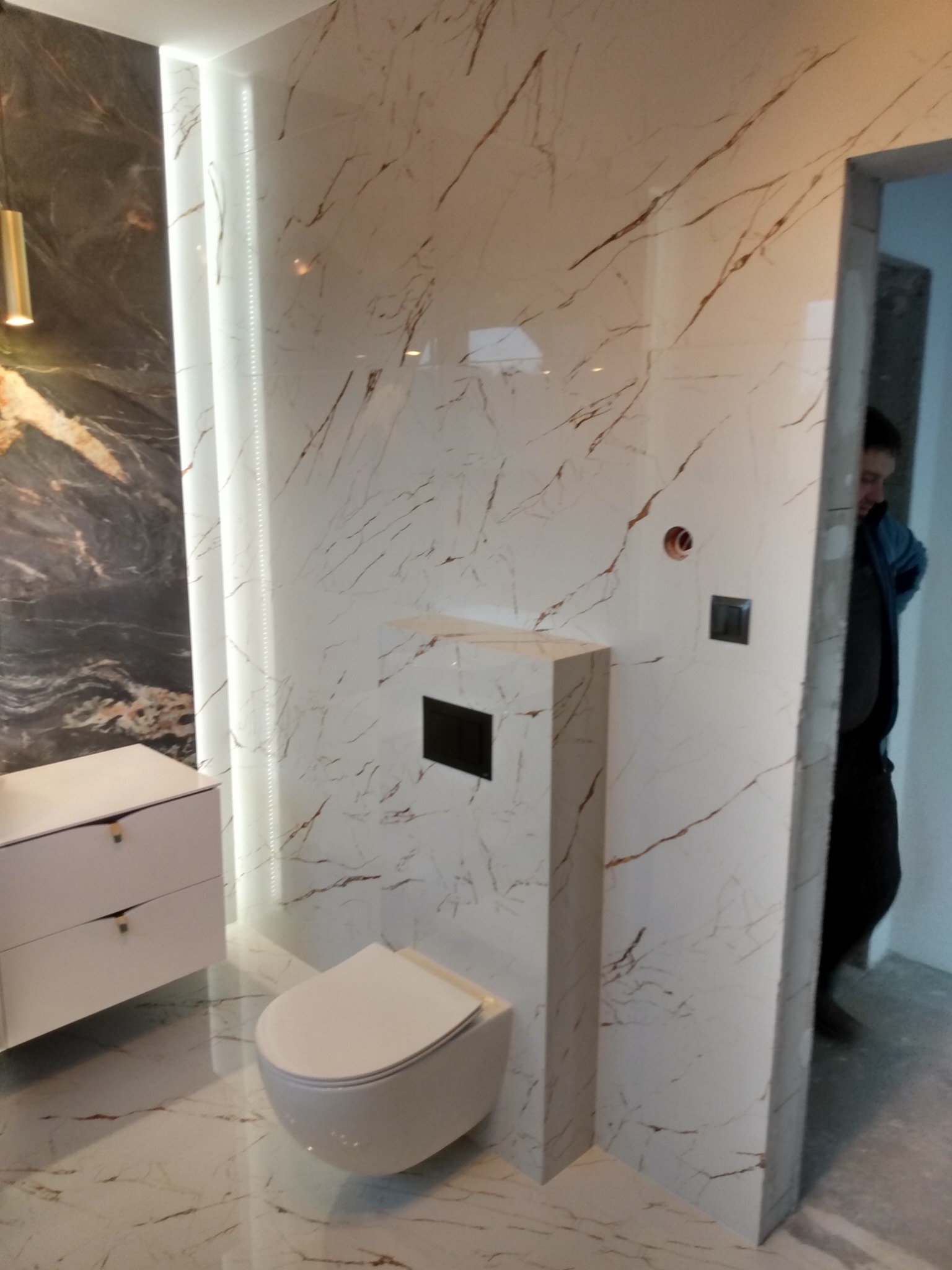 Nowoczesna łazienka z białymi płytkami imitującymi marmur z brązowymi żyłkami, podwieszana toaleta i biała szafka z szufladami, oświetlenie LED w pionowej wnęce, widoczny fragment osoby w przejściu.