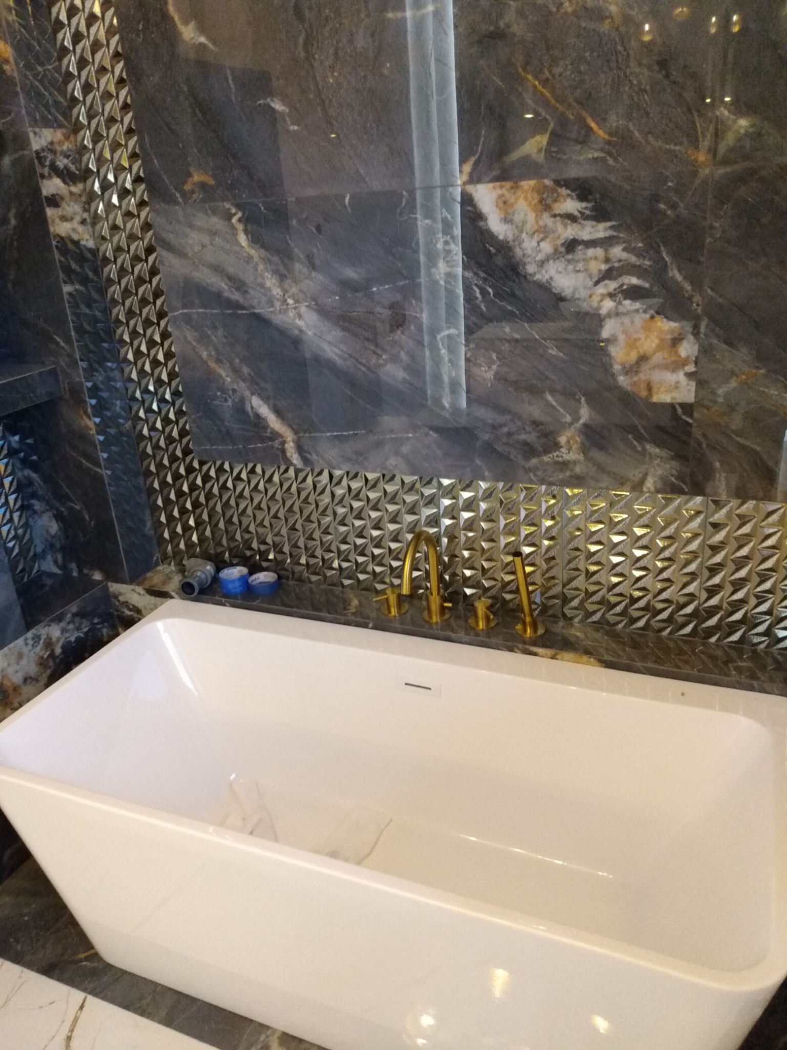 Biała, prostokątna wanna wolnostojąca z złotą armaturą na tle ściany wyłożonej ciemnym marmurem z dekoracyjnym, trójwymiarowym pasem płytek. Widoczne narzędzia instalacyjne na blacie.