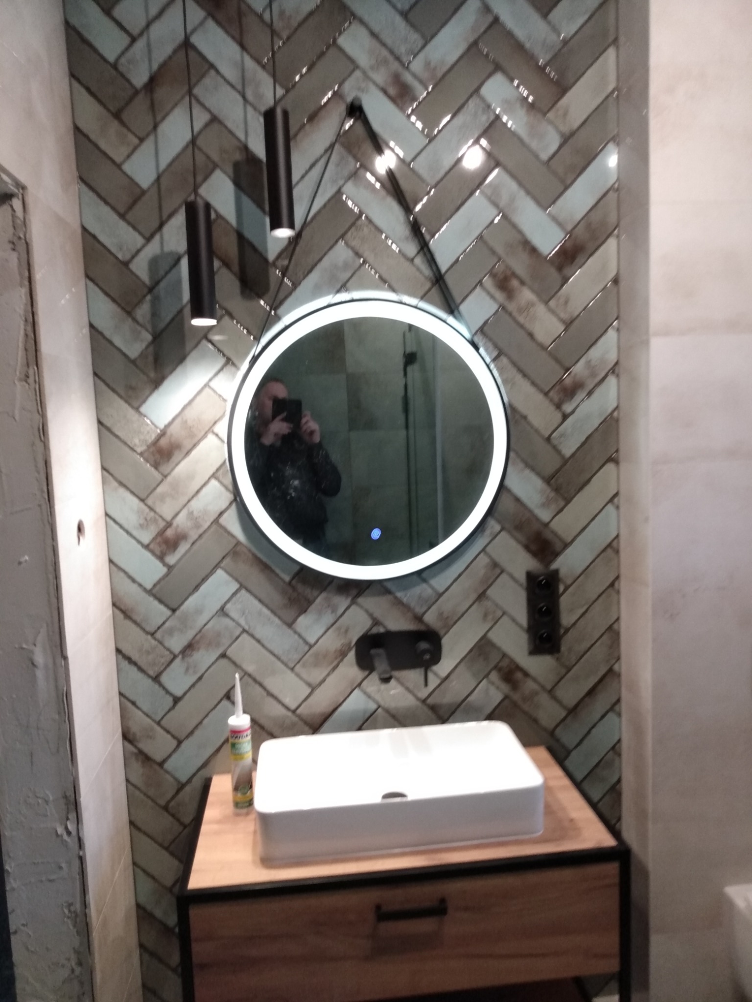 Ukończona łazienka z umywalką nablatową, lustrem z podświetleniem LED i wzorem jodełki na ścianie z płytek, widoczne lampy wiszące i silikon sanitarny.