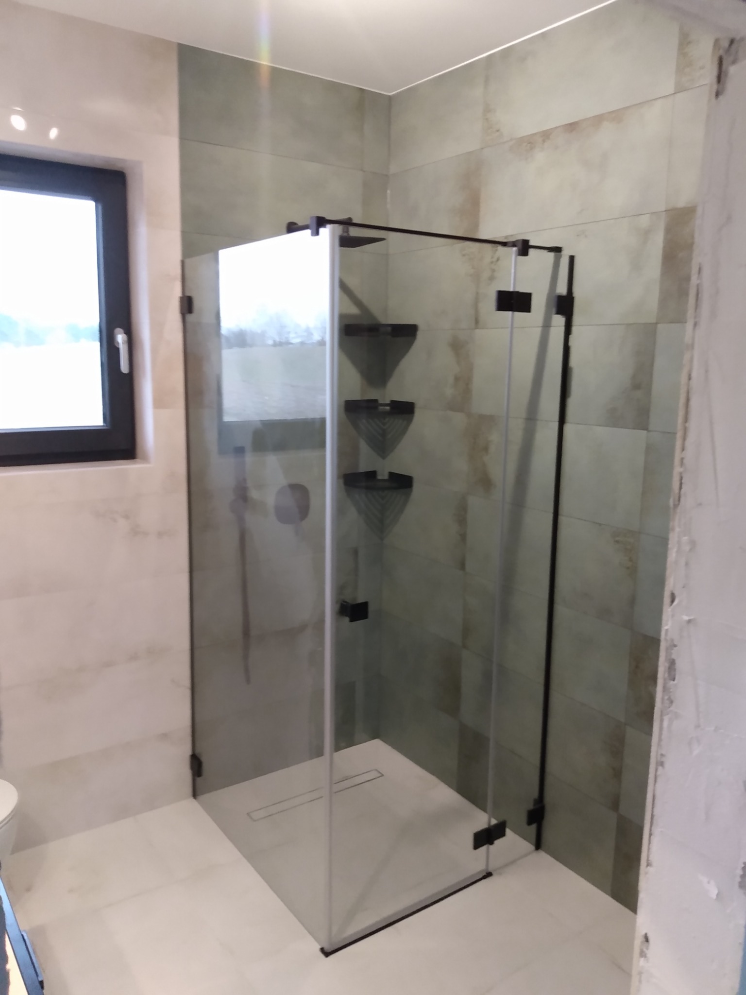 Nowoczesna kabina prysznicowa z czarnymi profilami i odpływem liniowym w Męcikale, wykończona płytkami w odcieniach beżu i szarości, widok z boku, okno w tle.