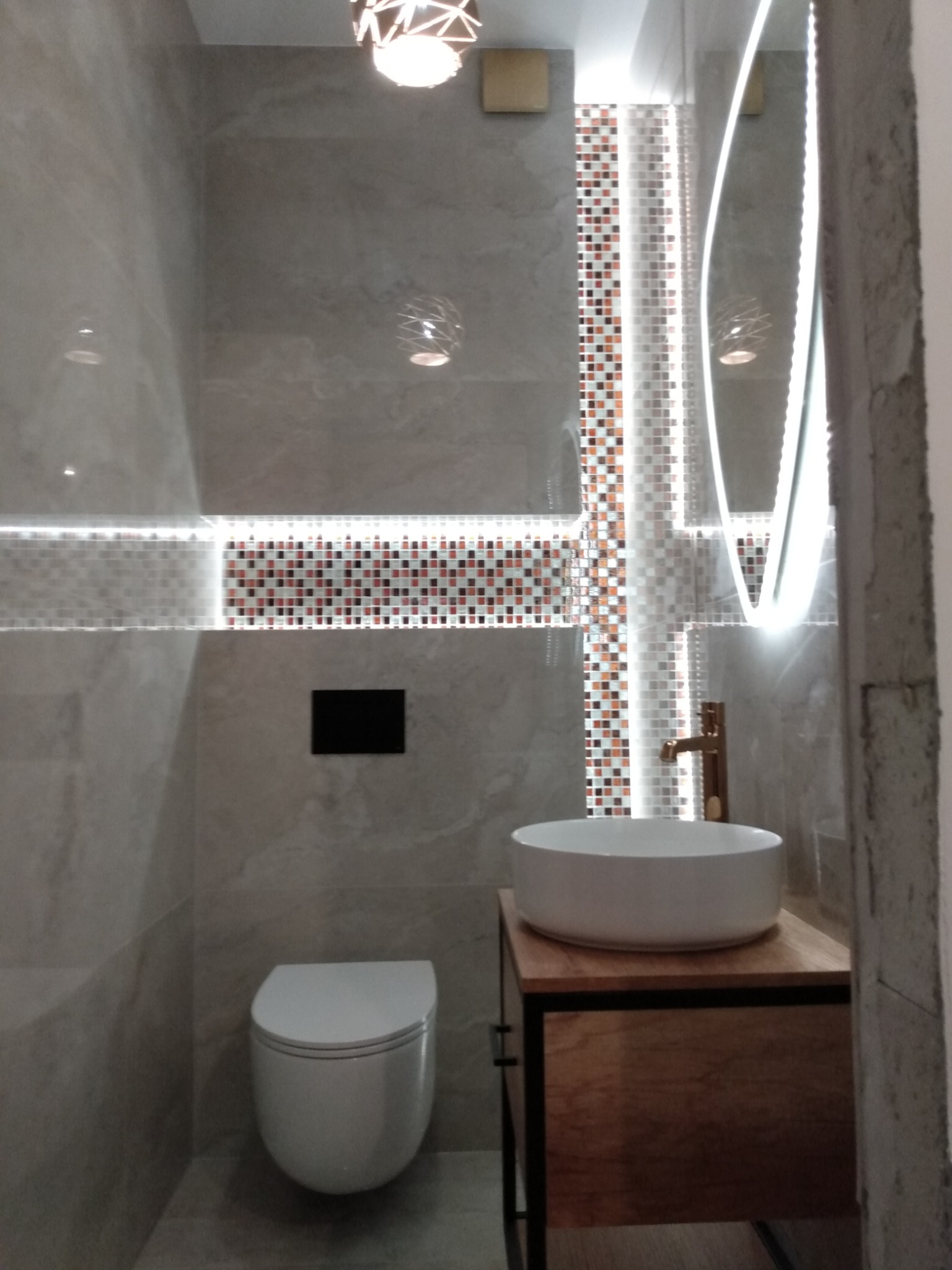 Nowoczesna łazienka z szarymi płytkami imitującymi marmur, mozaiką w odcieniach brązu i bieli, podświetlanym lustrem o nieregularnym kształcie, wiszącą miską WC i umywalką nablatową na drewnianej...