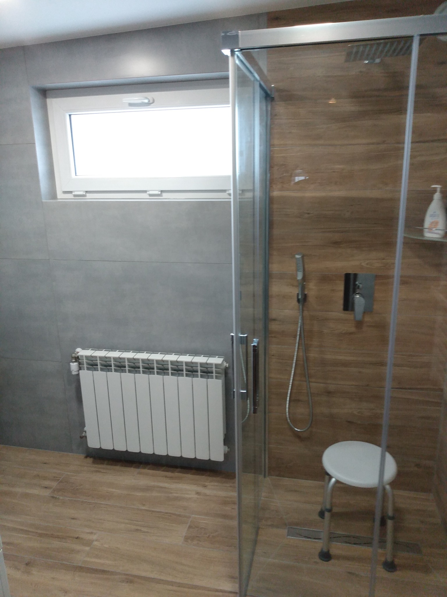 Nowoczesna łazienka z kabiną prysznicową typu walk-in, wyłożona szarymi płytkami na ścianach i drewnopodobnymi płytkami na podłodze oraz wewnątrz kabiny. Biały grzejnik pod oknem i małe siedzisko...