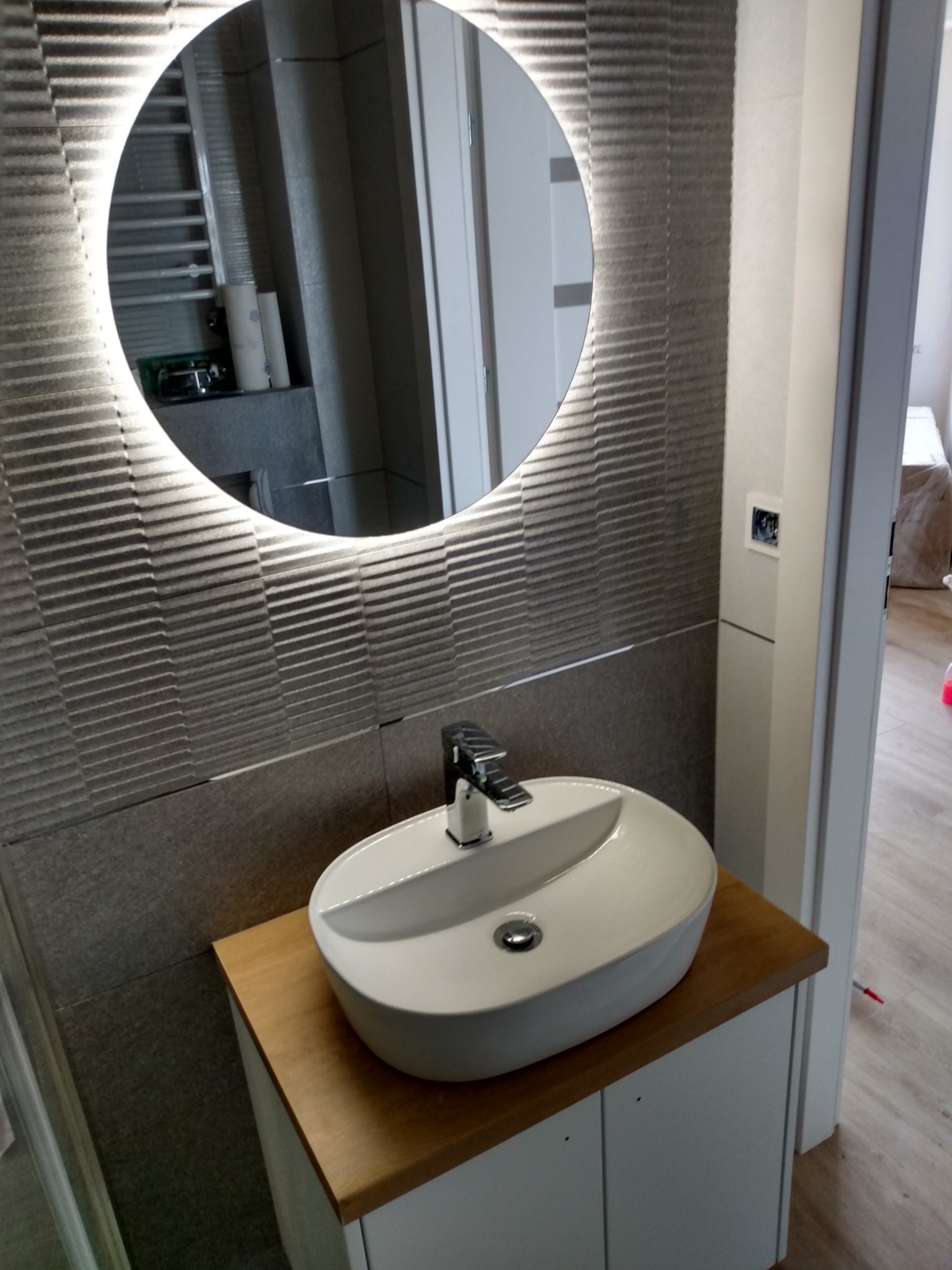 Nowoczesna łazienka z umywalką nablatową i okrągłym lustrem z podświetleniem LED, ściana wyłożona płytkami imitującymi fale.