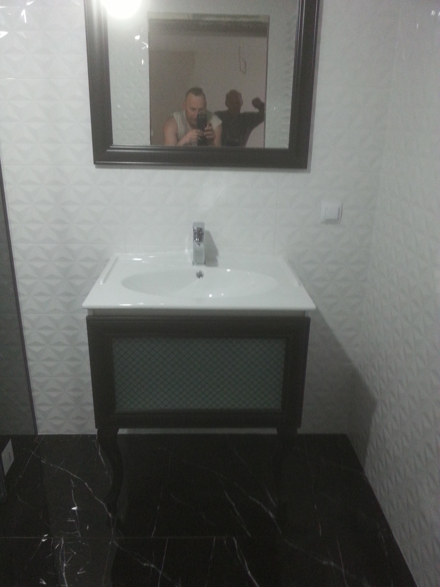 Nowoczesna łazienka z czarną marmurową podłogą i białą umywalką z szafką w ciemnej ramie; w lustrze odbija się osoba robiąca zdjęcie i druga osoba.