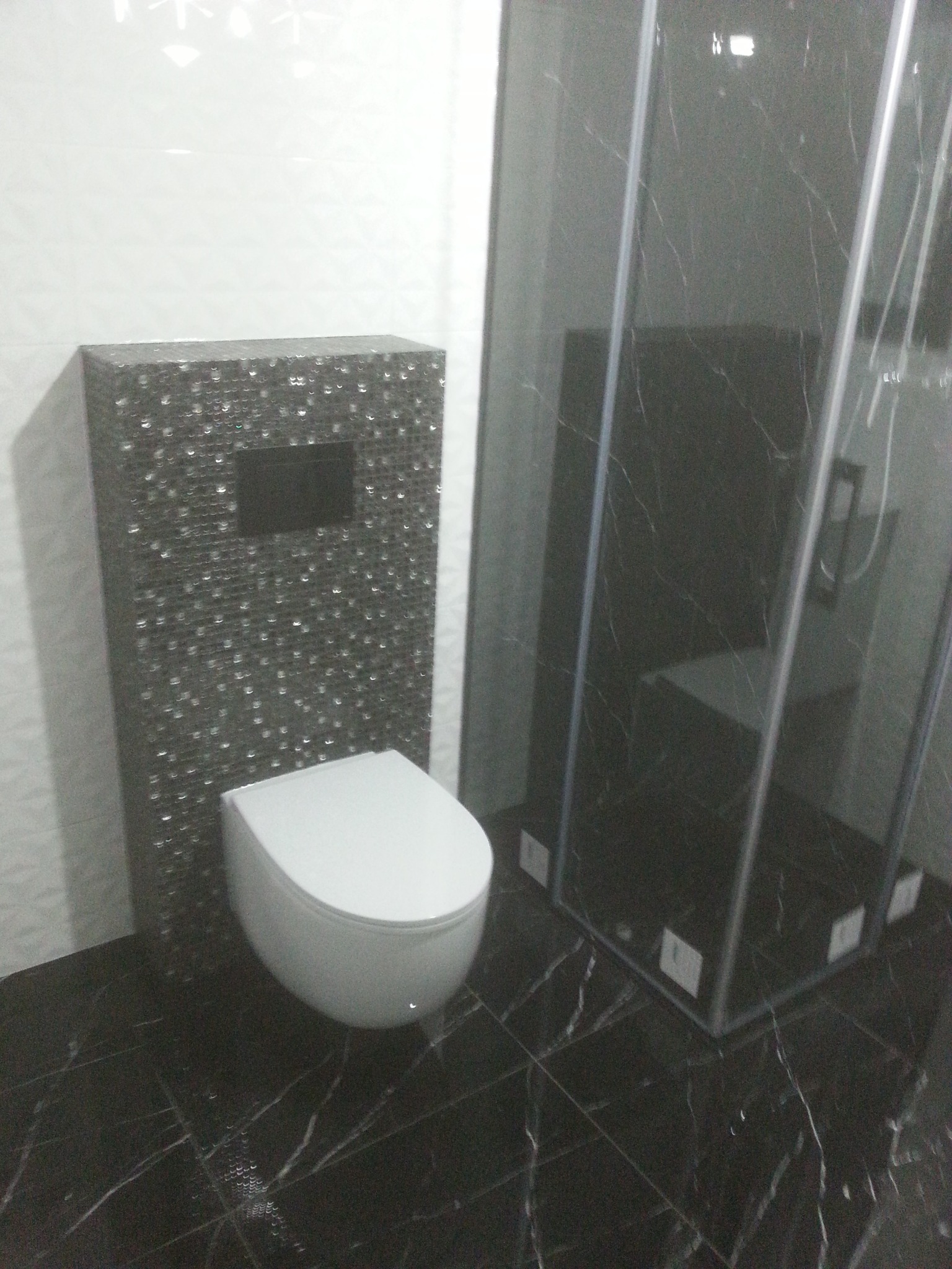 Nowoczesna łazienka z czarną marmurową podłogą i kabiną prysznicową, toaleta podwieszana na tle ściany z mozaiki.