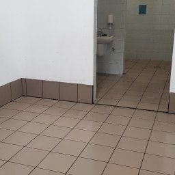 Dawid Pielok - Wnętrze pomieszczenia z beżowymi płytkami podłogowymi i brązowymi cokołami, widoczna umywalka i toaleta w tle, ściany białe z widocznymi niedoskonałościami.