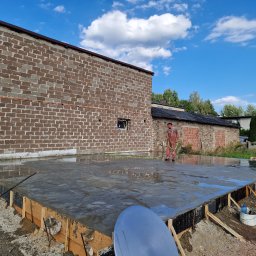 Dawid Pielok - Pracownik w czerwonym kombinezonie polewa wodą świeżo wylany, mokry beton na przygotowanym fundamencie budynku z cegły.