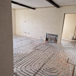 Dawid Pielok - Instalacja ogrzewania podłogowego z białych rur na folii z czerwoną kratką, widoczne kolektory w ścianie i surowy stan ścian z elementami konstrukcyjnymi.