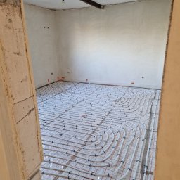 Dawid Pielok - Instalacja ogrzewania podłogowego w surowym stanie mieszkania, widok zza framugi drzwi, widoczne rury na folii z siatką, ściany przygotowane do dalszych prac.
