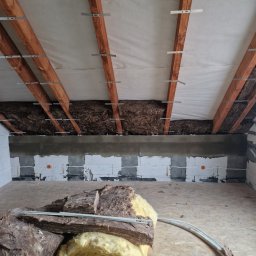 Dawid Pielok - Wnętrze poddasza w trakcie ocieplania: widoczne drewniane belki stropowe z metalowymi mocowaniami, częściowo ułożona izolacja z wełny mineralnej oraz fragment ściany z instalacją elektryczną.