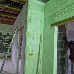 Dawid Pielok - Wnętrze w trakcie prac wykończeniowych: ściany z betonu komórkowego z instalacją elektryczną, częściowo pokryte gładzią, drabina oparta o ścianę, widoczne drewniane elementy konstrukcyjne stropu.