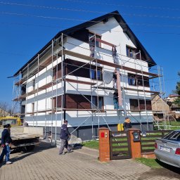 Dawid Pielok - Dwupiętrowy dom z ciemnymi drewnianymi elementami na elewacji w trakcie prac na rusztowaniu, kilku pracowników przenosi deski, na podjeździe stoi samochód i przyczepka.