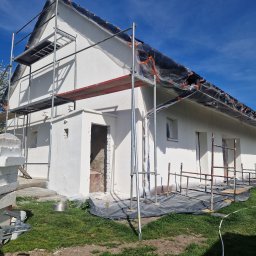 Dawid Pielok - Dom jednorodzinny w trakcie prac elewacyjnych, świeżo otynkowany na biało, z rozstawionym rusztowaniem i folią ochronną na dachu, widok na fragment niezabezpieczonego wejścia i stos styropianu.