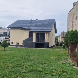 Dawid Pielok - Widok zewnętrzny domu z beżową elewacją i ciemnoszarą stolarką okienną oraz dachem z blachodachówki, otoczonego zielonym trawnikiem.