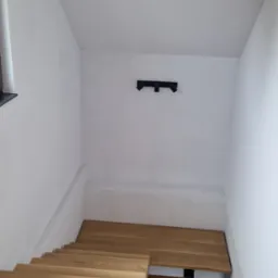 Klatka schodowa z drewnianymi schodami i białymi ścianami, widok z góry, z zamontowanym oświetleniem sufitowym na ścianie.