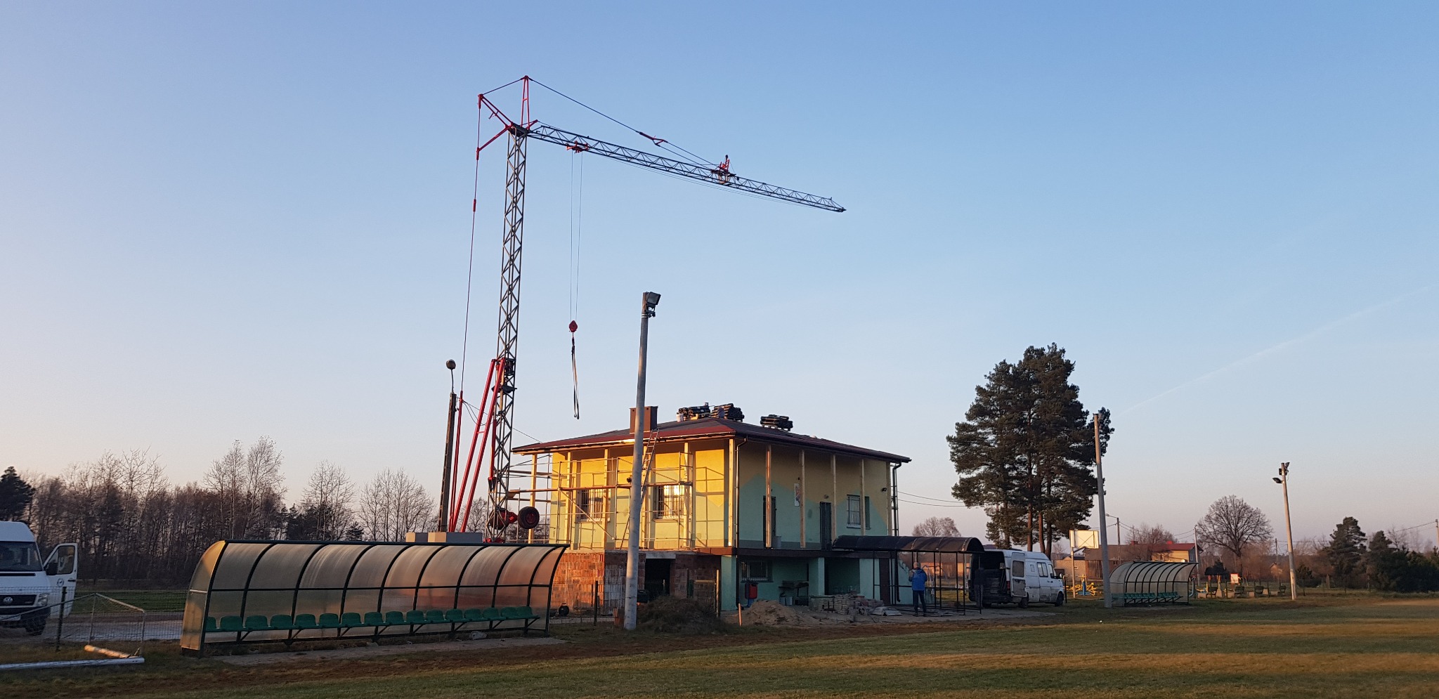 Budynek w trakcie remontu elewacji, widoczny dźwig budowlany, rusztowania i częściowo ocieplona ściana, na pierwszym planie wiata dla zawodników.