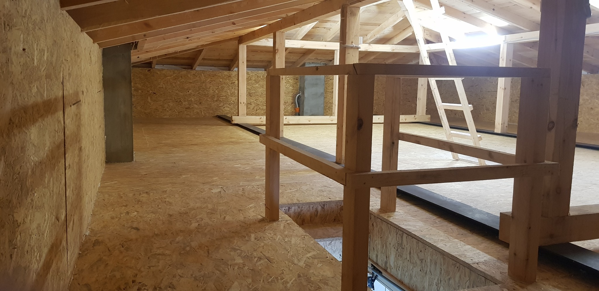 Poddasze w trakcie adaptacji z widoczną konstrukcją drewnianą dachu, podłogą z płyt OSB i fragmentem betonowego filaru.