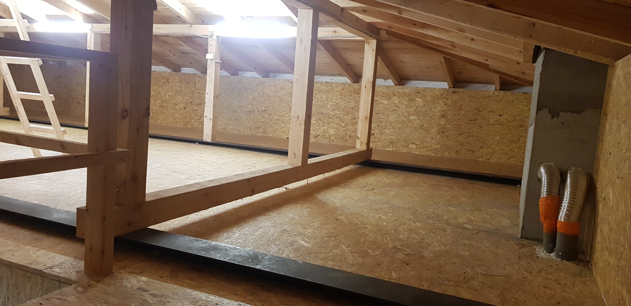 Poddasze w trakcie adaptacji: drewniana konstrukcja szkieletowa, płyty OSB na ścianach i podłodze, widoczne przewody wentylacyjne z pomarańczową taśmą.