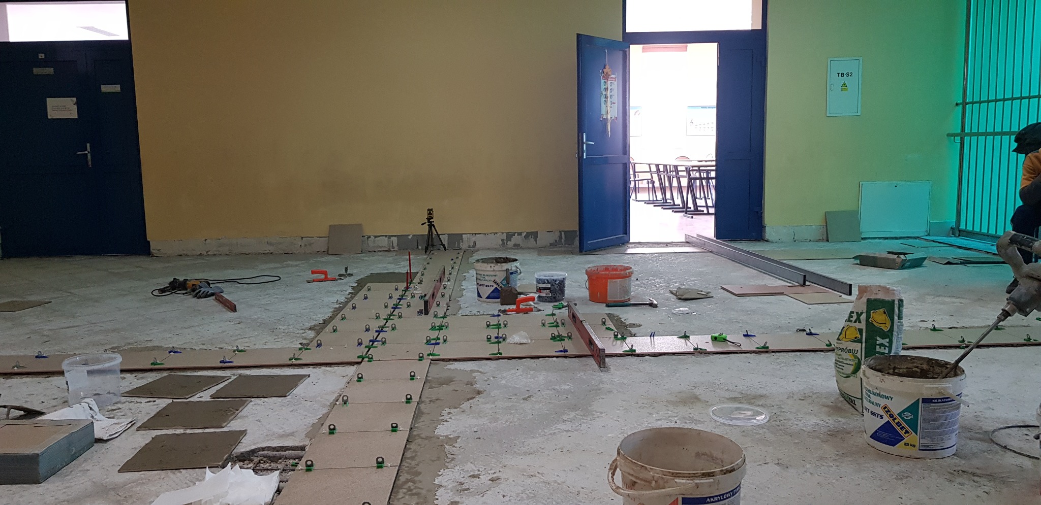 Układanie płytek podłogowych w pomieszczeniu, widoczne płytki rozłożone wzdłuż linii, poziomica, wiadro z klejem, worek z zaprawą klejową, oraz osoba mieszająca zaprawę mieszadłem elektrycznym.