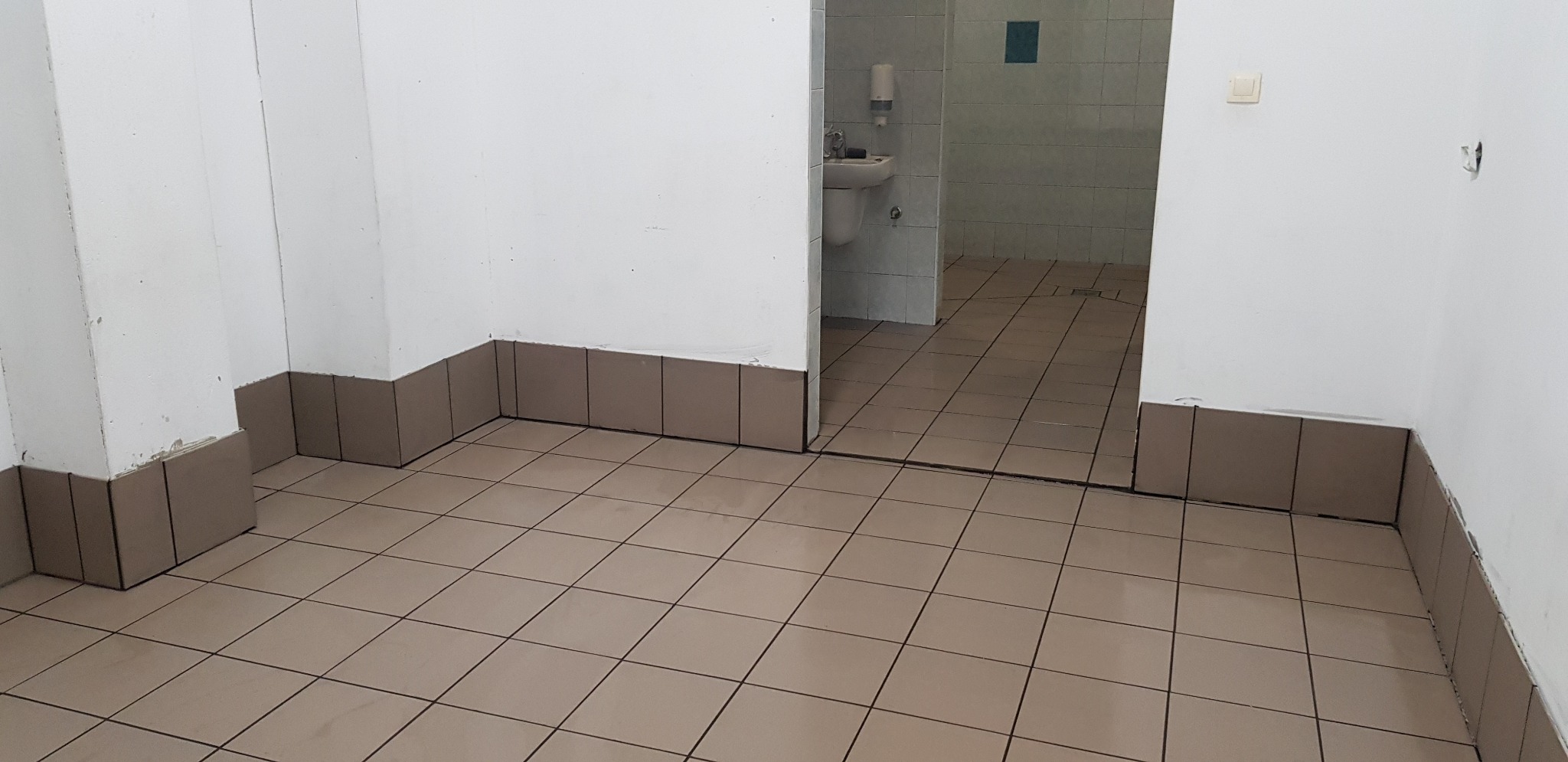 Wnętrze pomieszczenia z beżowymi płytkami podłogowymi i brązowymi cokołami, widoczna umywalka i toaleta w tle, ściany białe z widocznymi niedoskonałościami.