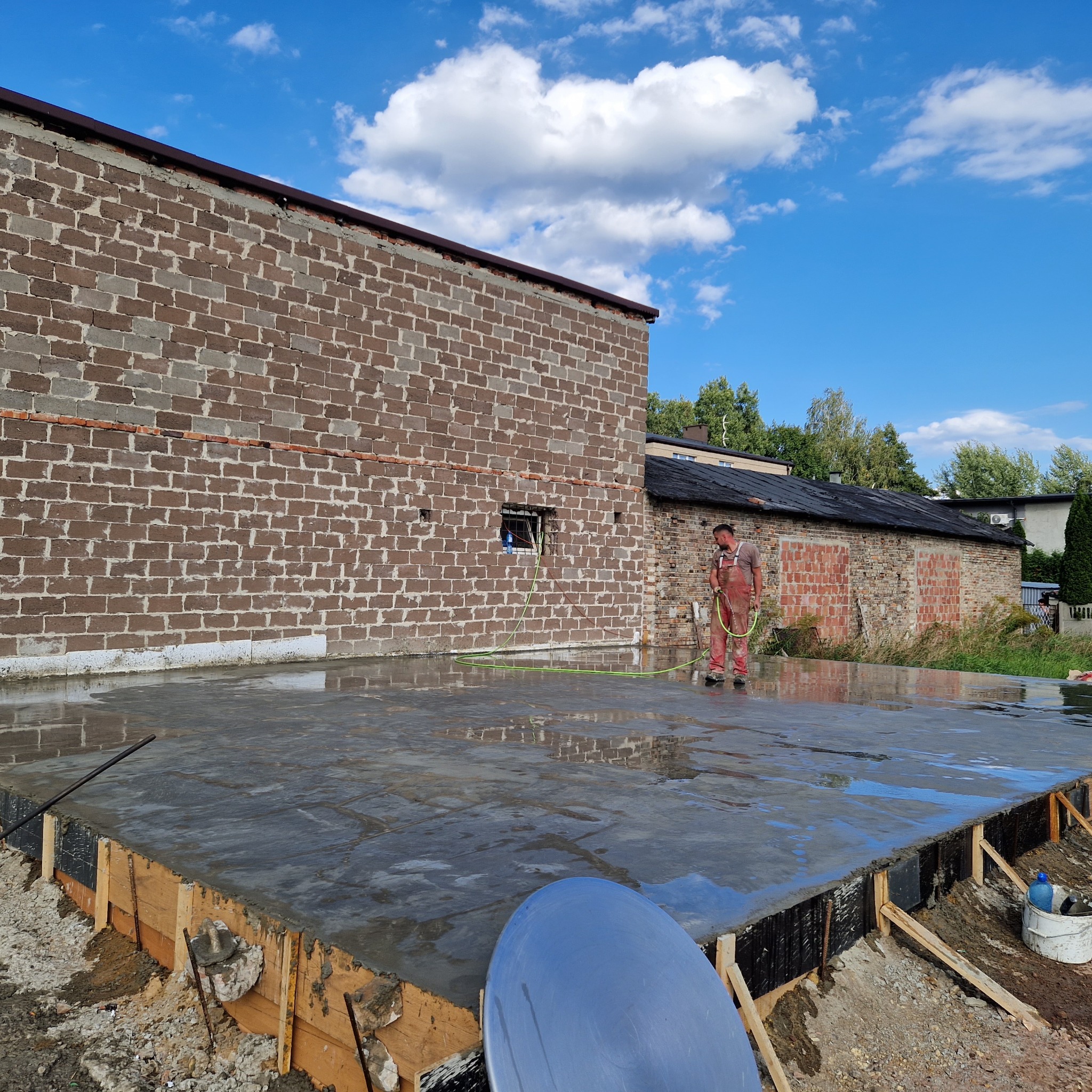 Pracownik w czerwonym kombinezonie polewa wodą świeżo wylany, mokry beton na przygotowanym fundamencie budynku z cegły.