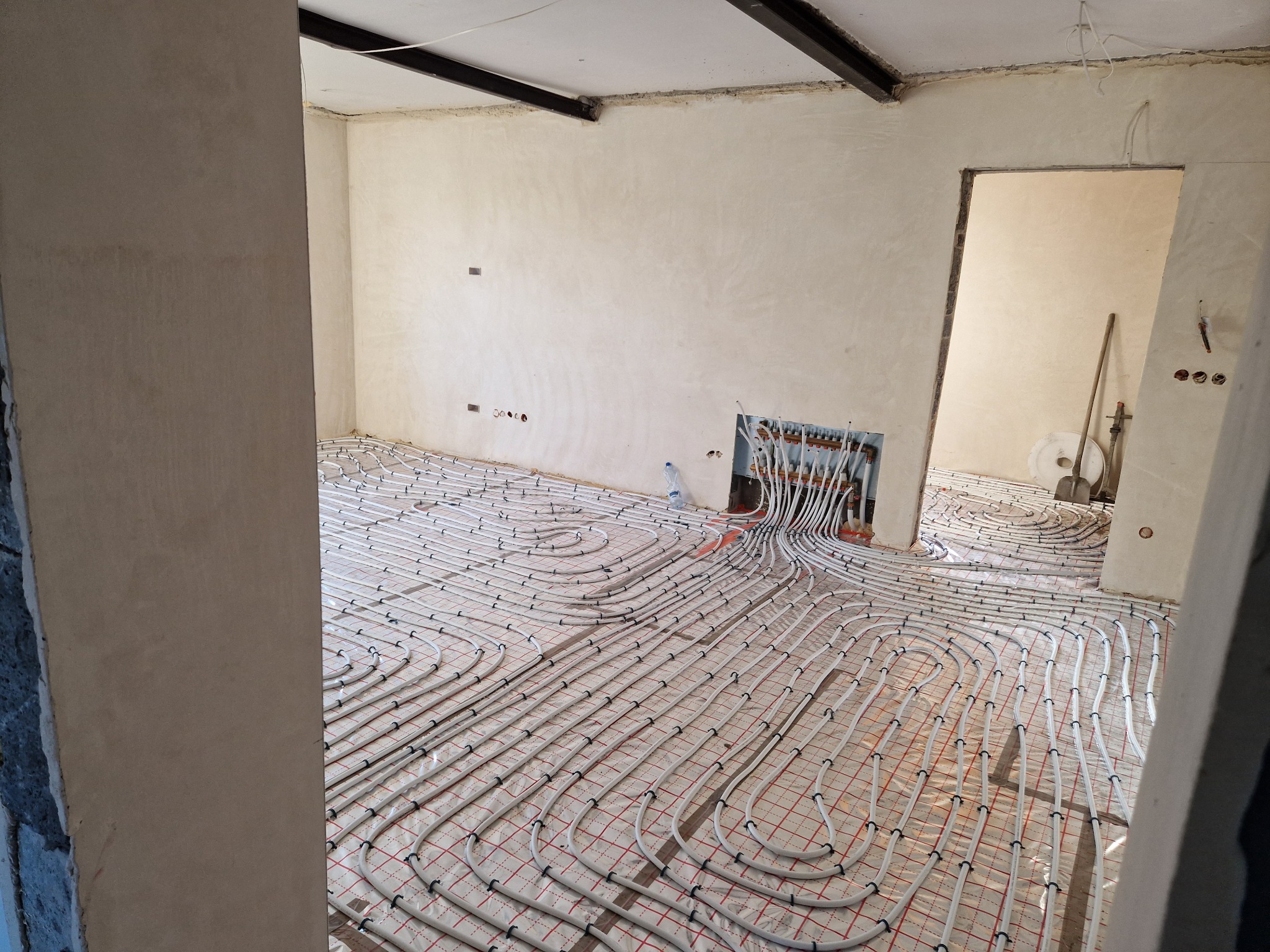Instalacja ogrzewania podłogowego z białych rur na folii z czerwoną kratką, widoczne kolektory w ścianie i surowy stan ścian z elementami konstrukcyjnymi.