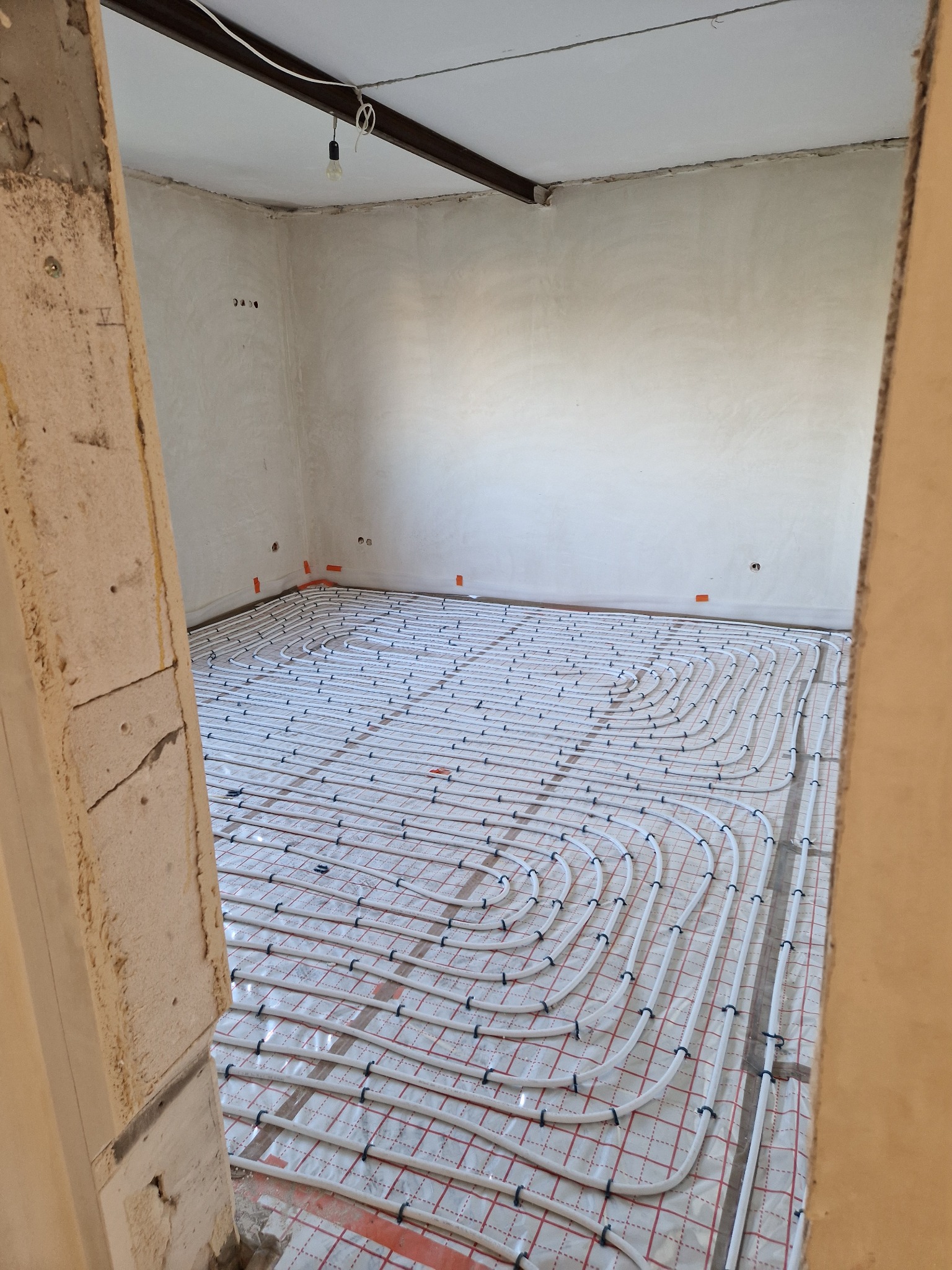 Instalacja ogrzewania podłogowego w surowym stanie mieszkania, widok zza framugi drzwi, widoczne rury na folii z siatką, ściany przygotowane do dalszych prac.