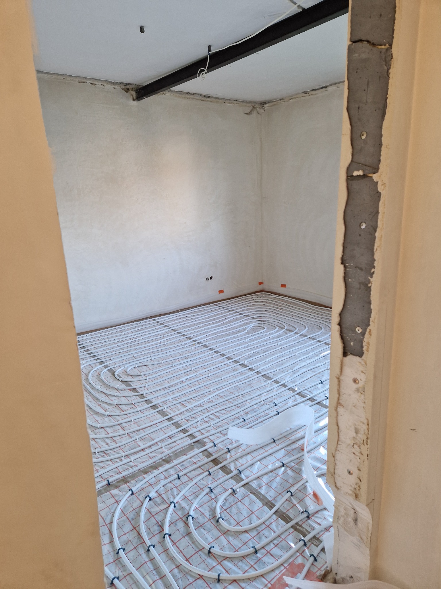 Instalacja ogrzewania podłogowego w surowym pomieszczeniu z gładzią na ścianach, widok przez niedokończone futryny drzwi.