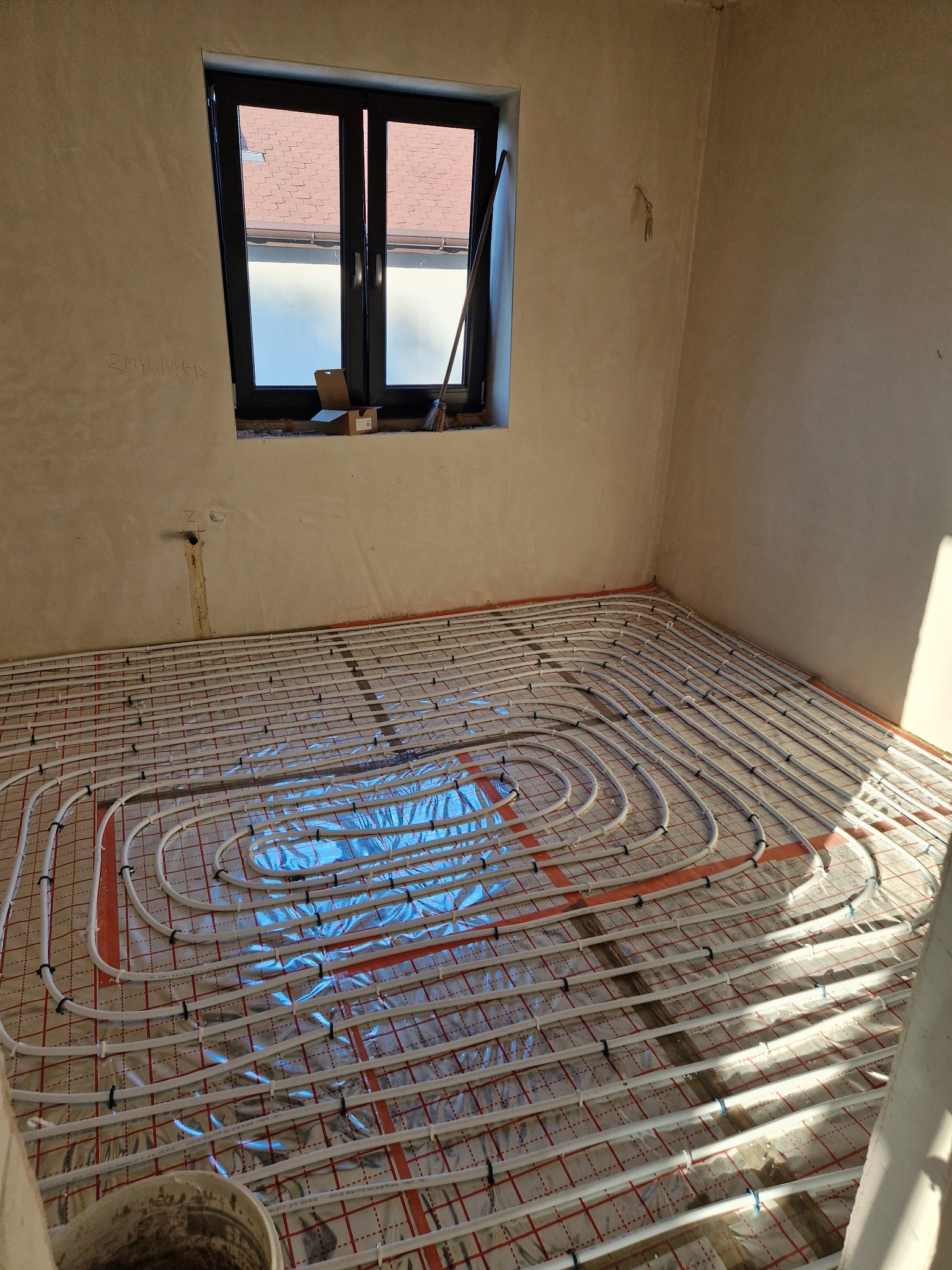 Białe rury ogrzewania podłogowego rozłożone na folii izolacyjnej w surowym wnętrzu pokoju z oknem, widoczny fragment ściany z napisem 'ZMYWACKA'.