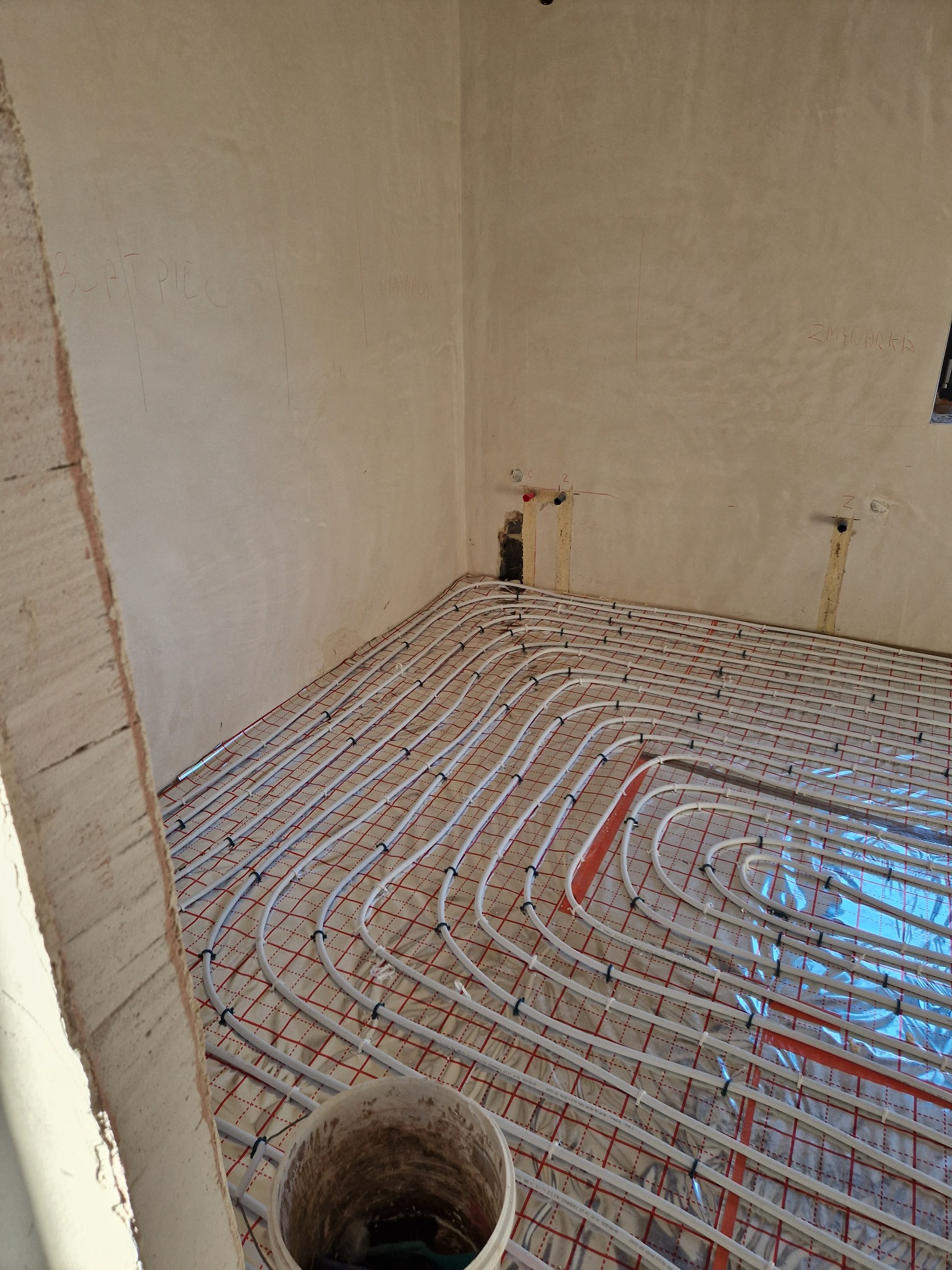 Instalacja ogrzewania podłogowego w surowym wnętrzu, widoczne rury grzewcze na folii z czerwoną kratką, ściany z napisami wykonanymi ołówkiem, wiadro w rogu pomieszczenia.
