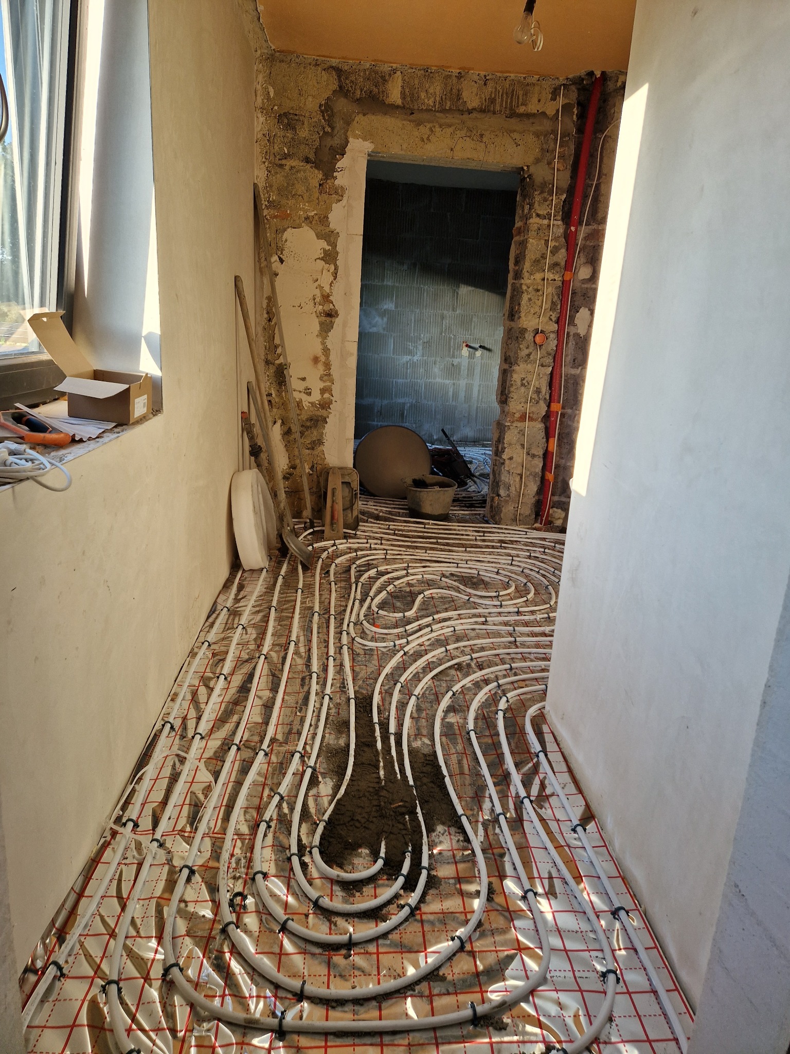 Instalacja ogrzewania podłogowego w trakcie prac, widoczne rury na folii z siatką, narzędzia w tle, surowe ściany w trakcie remontu.