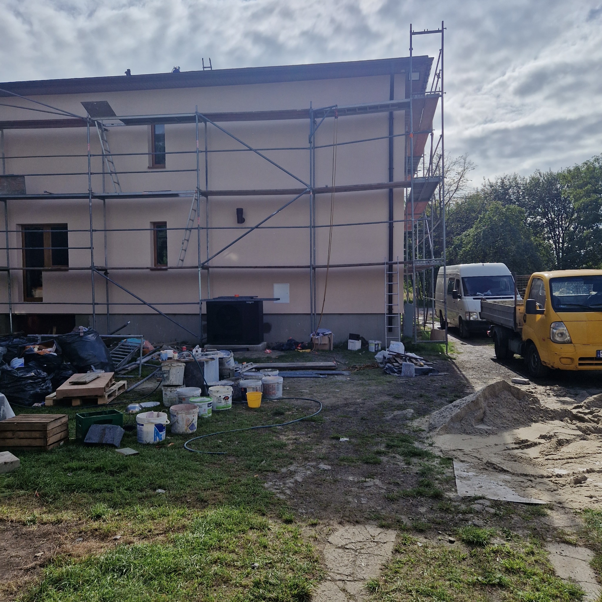 Dwupiętrowy dom w trakcie prac elewacyjnych, otoczony rusztowaniem, z widocznymi wiadrami farby i dwoma samochodami dostawczymi na podwórku.