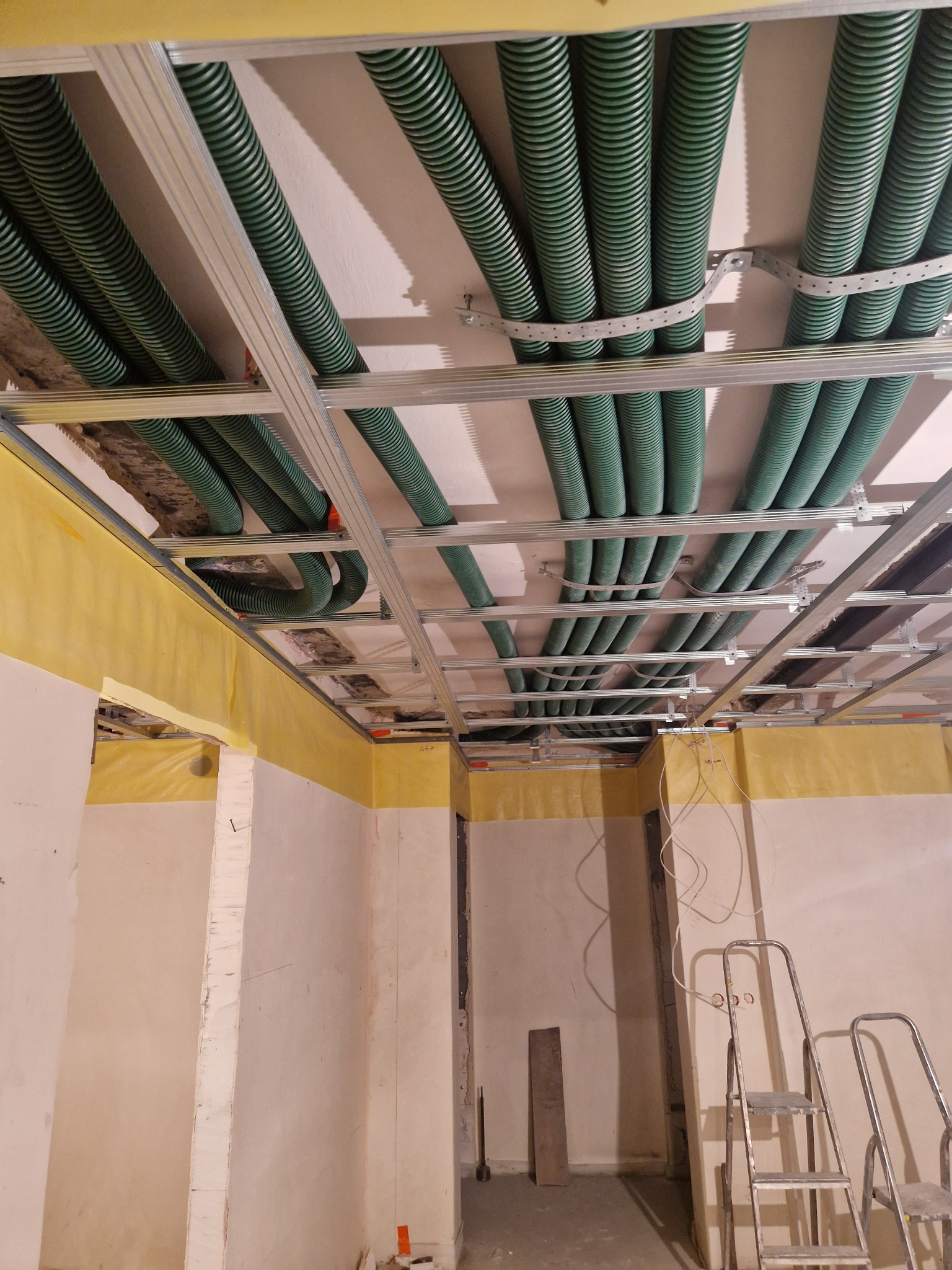 Instalacja zielonych rur karbowanych podwieszonych do metalowej konstrukcji sufitu w trakcie prac wykończeniowych w pomieszczeniu.