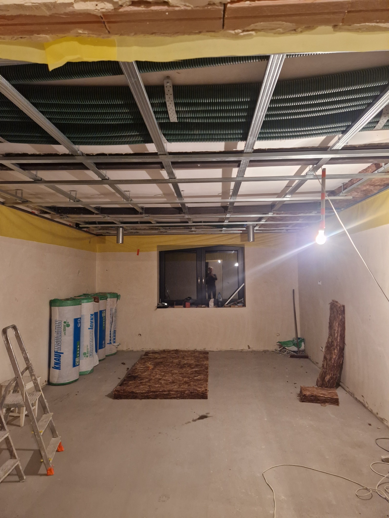 Wnętrze w trakcie montażu sufitu podwieszanego z widoczną konstrukcją metalową, izolacją termiczną w rolkach Knauf Insulation, częściowo ułożoną wełną mineralną na podłodze i lampą wiszącą na kablu.