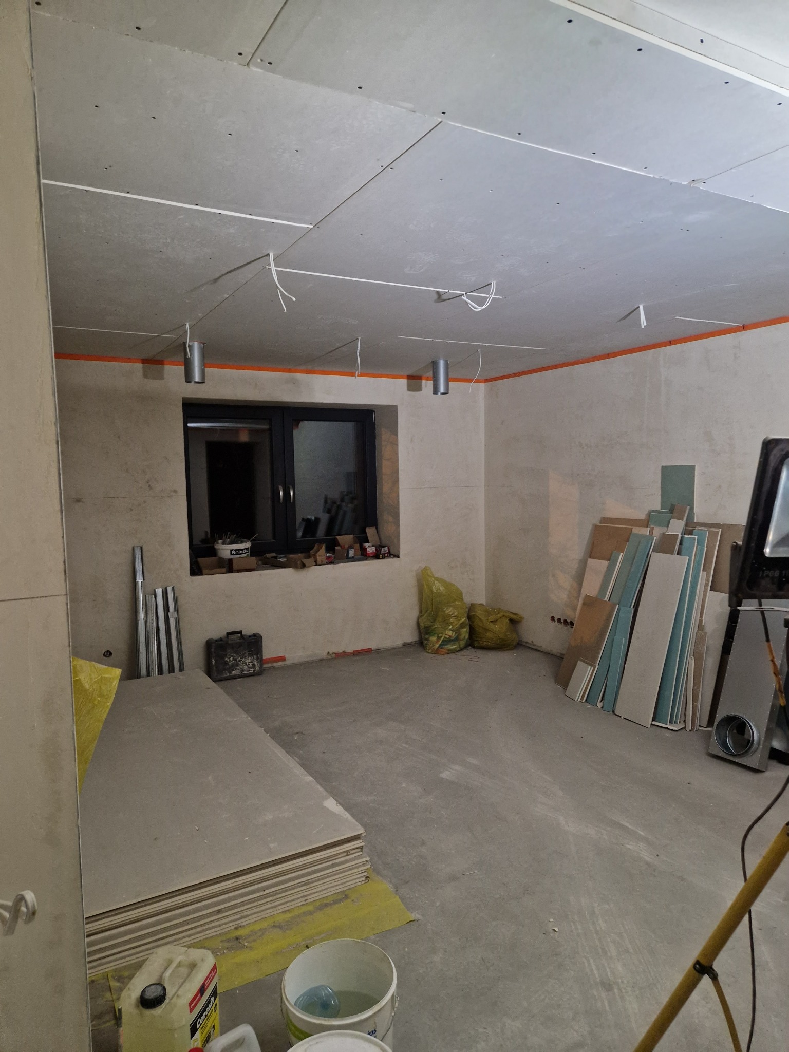 Wnętrze w trakcie remontu: sufit z płyt gipsowo-kartonowych z widocznymi łączeniami, przewody elektryczne, ściany z pozostałościami gładzi, okno z narzędziami na parapecie, stos płyt gipsowych...