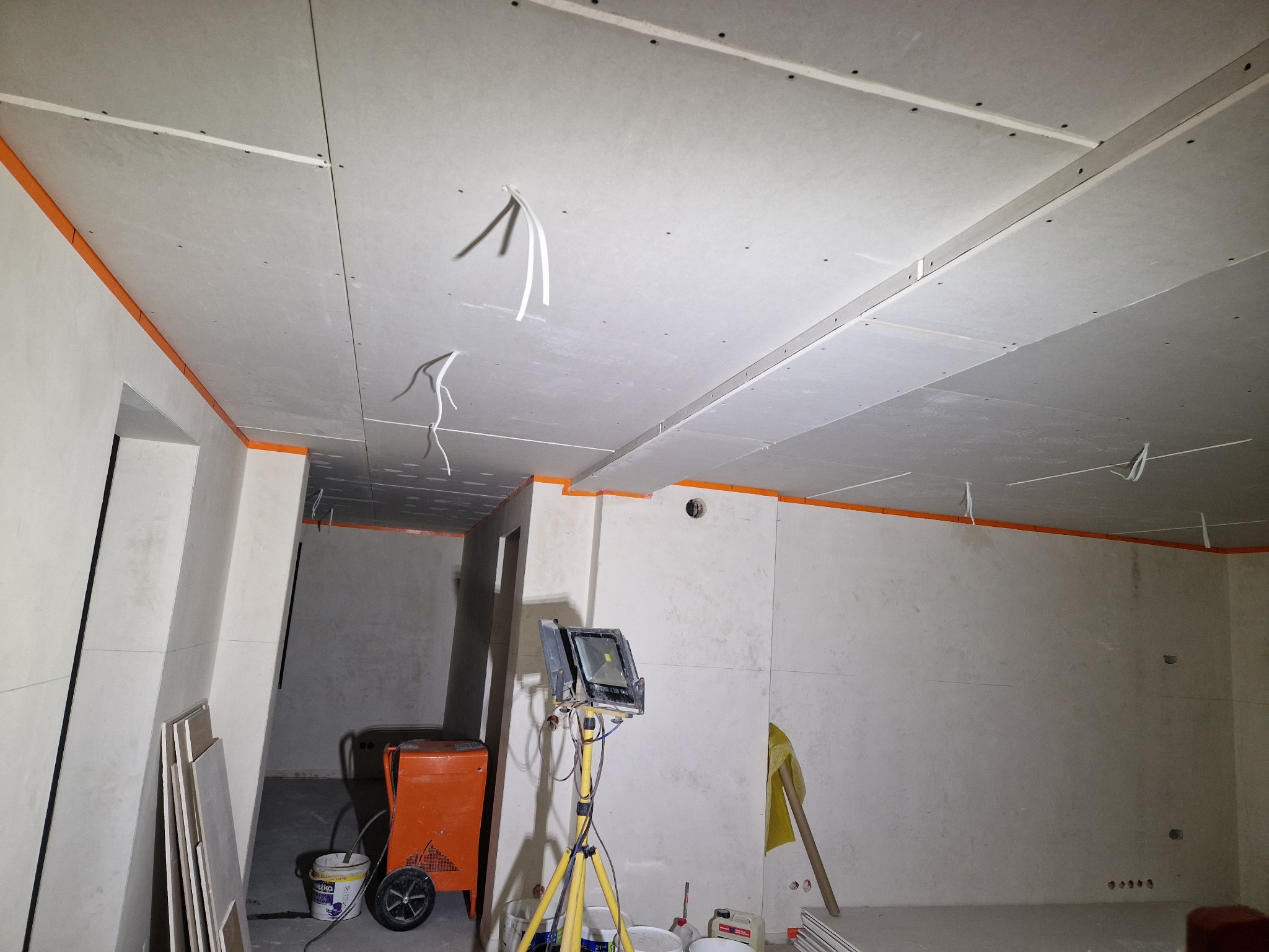Wnętrze w trakcie prac wykończeniowych: sufit i ściany pokryte płytami gipsowo-kartonowymi, widoczne kable, pomarańczowe taśmy zabezpieczające, przenośny reflektor na trójnogu, oraz pomarańczowy...