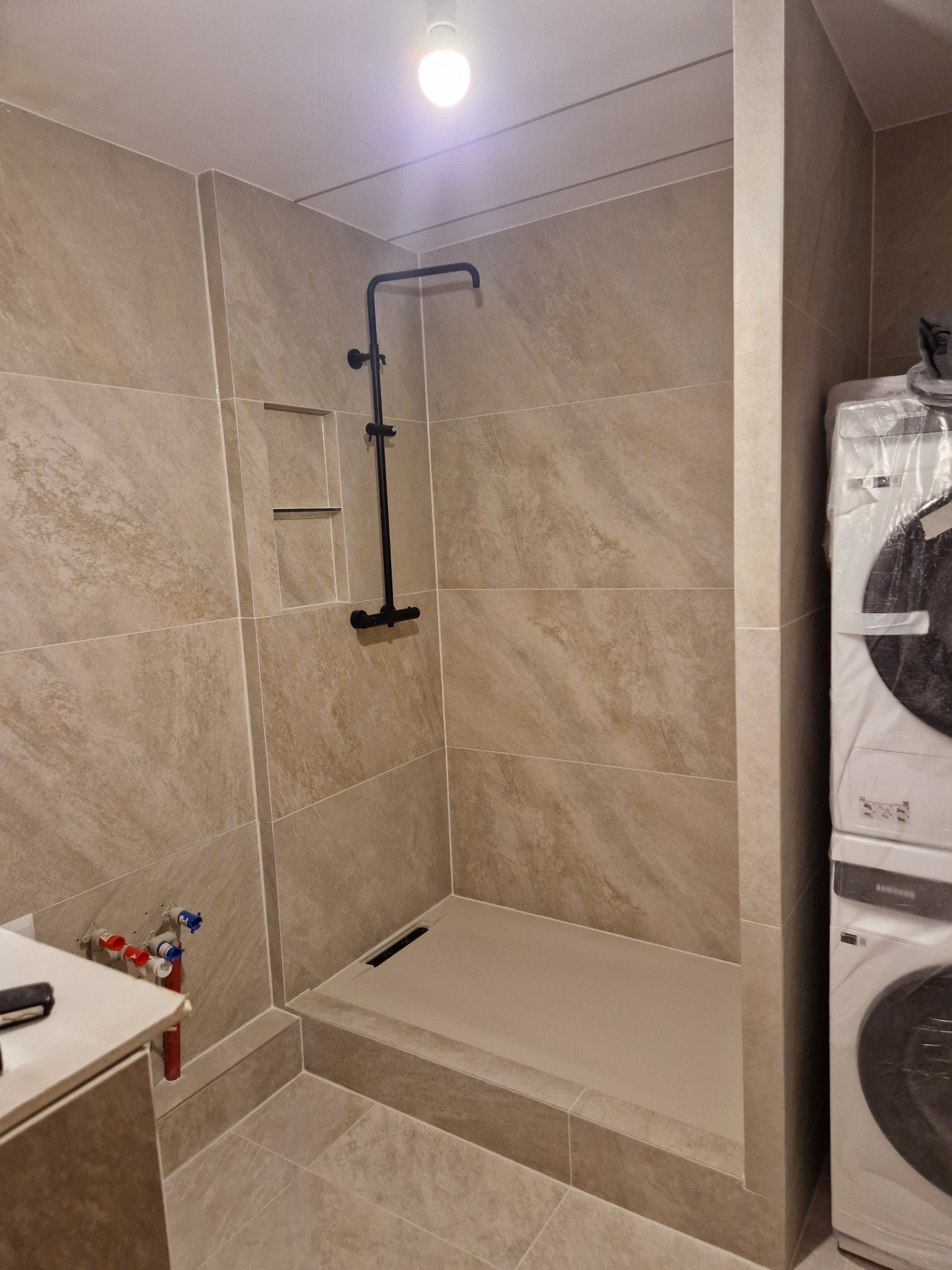 Nowoczesna kabina prysznicowa z beżowymi płytkami imitującymi kamień, czarną armaturą i wnęką na kosmetyki, częściowo widoczna pralka w folii ochronnej.