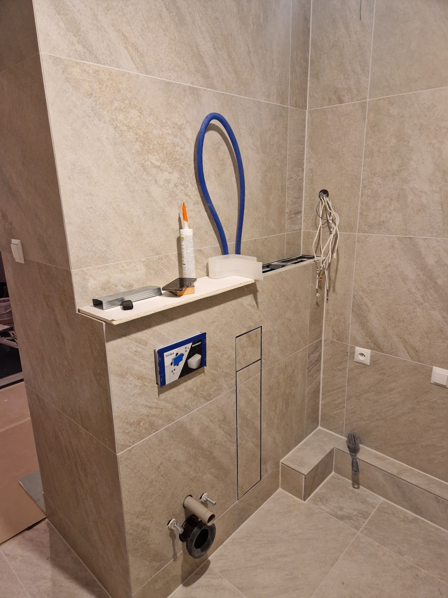 Montaż podtynkowego stelaża WC z widocznymi przyłączami wodno-kanalizacyjnymi, częściowo wykończony płytkami w odcieniach beżu, z akcesoriami montażowymi na półce i wystającymi przewodami...