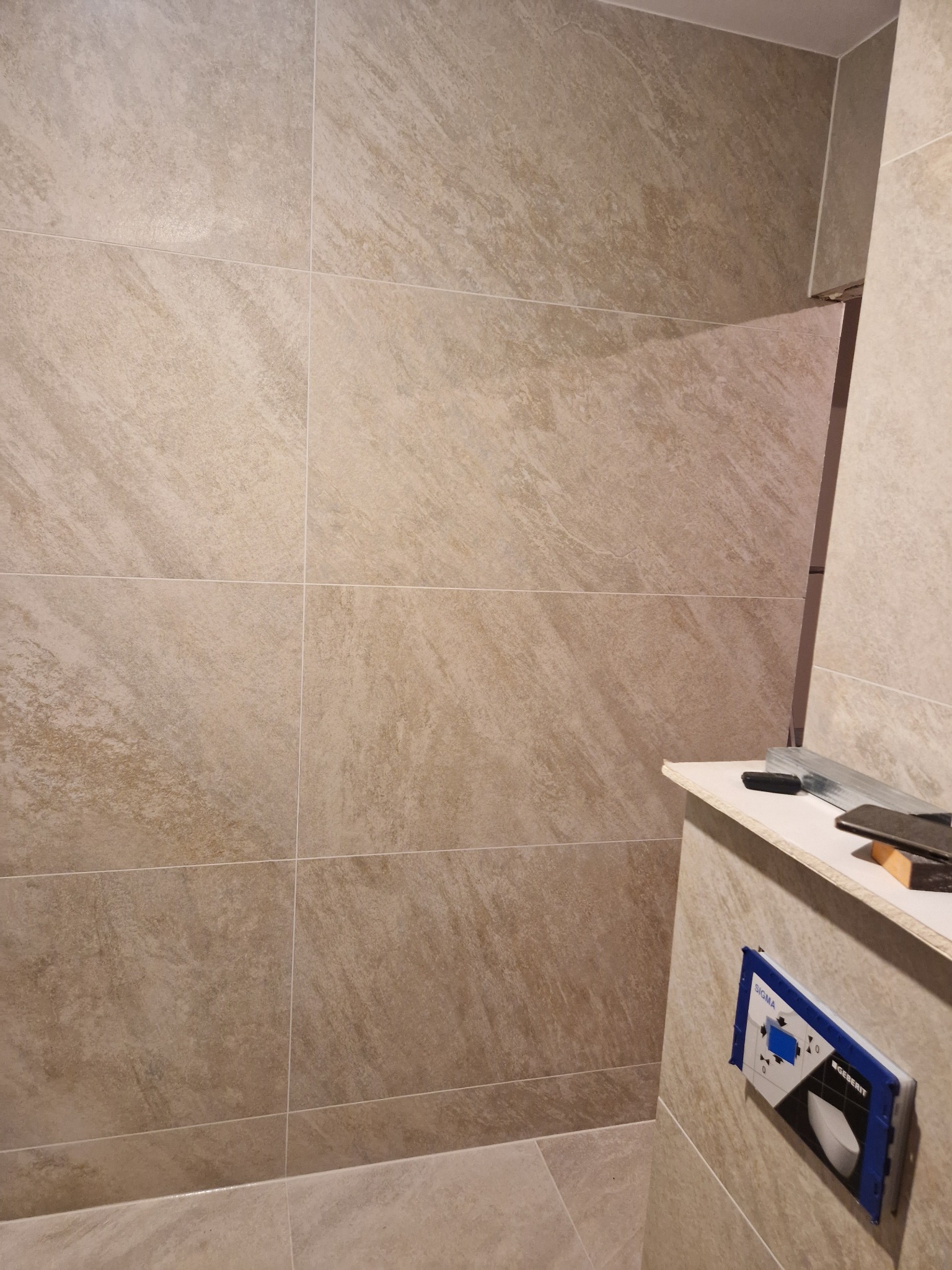 Beżowe płytki imitujące kamień na ścianie w łazience, widoczny fragment stelaża podtynkowego Geberit i narzędzia na półce, sugerujące prace wykończeniowe.